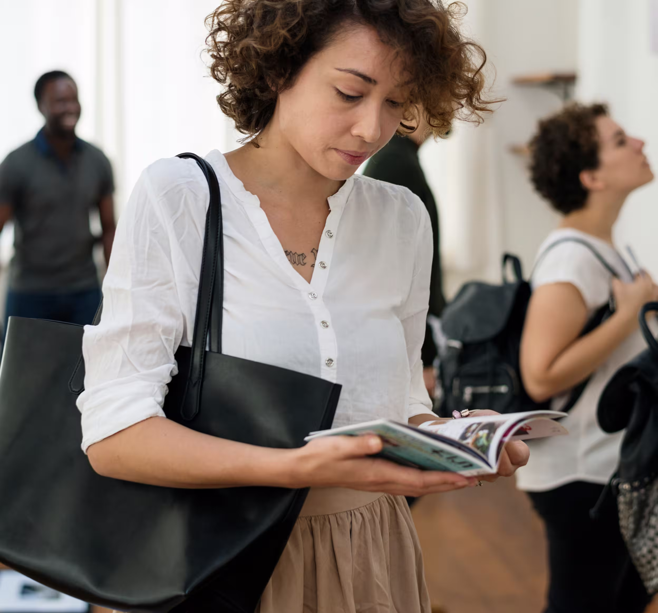 Une femme lisant un livret, sac noir à l'épaule, avec des personnes floues en arrière-plan.