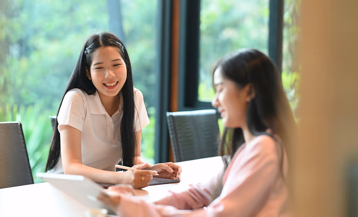 Deux femmes souriantes discutent à une table, l'une utilisant une tablette, devant une grande fenêtre.