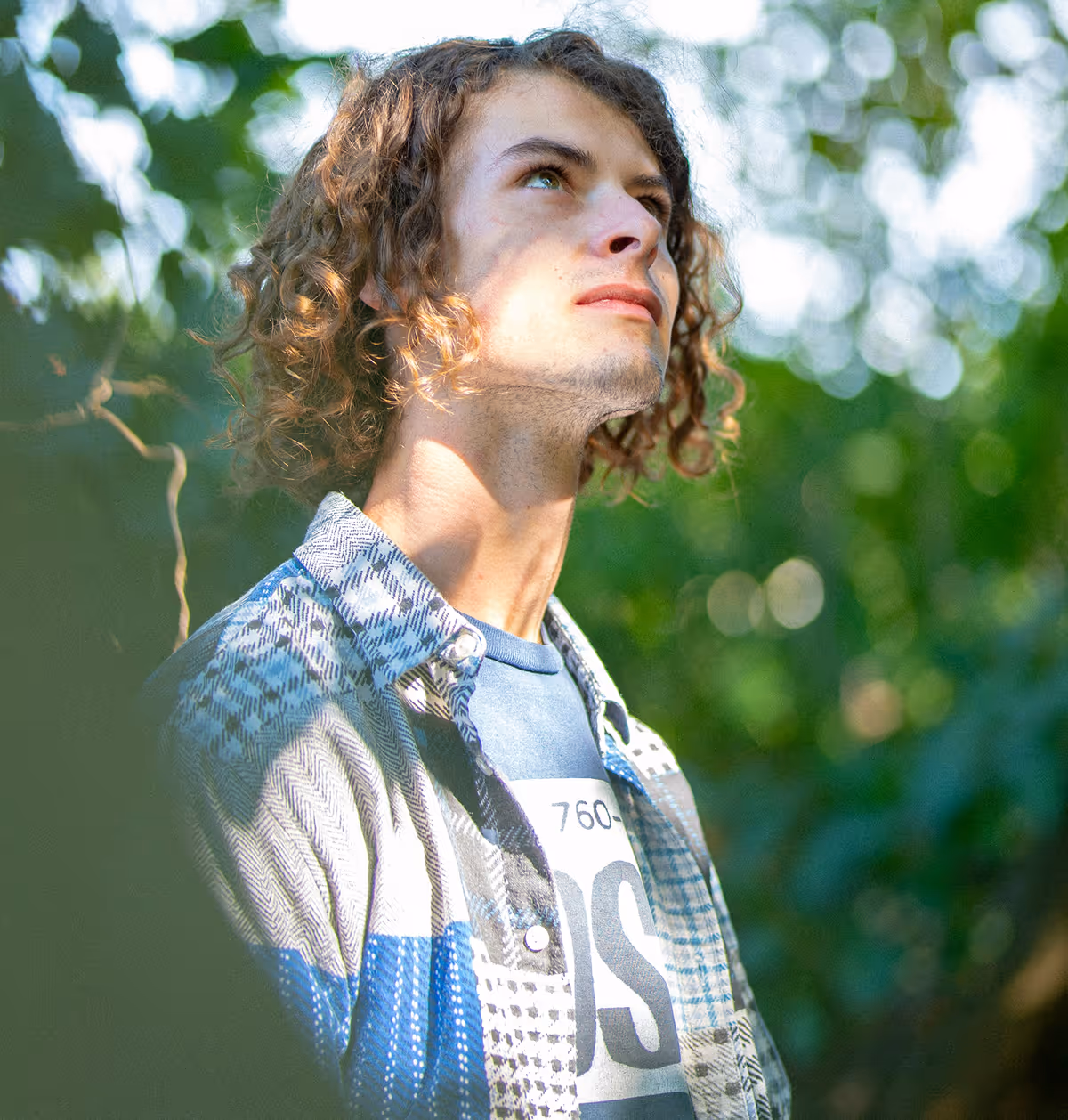 Jeune homme aux cheveux bouclés regardant vers le haut dans un environnement naturel ensoleillé.