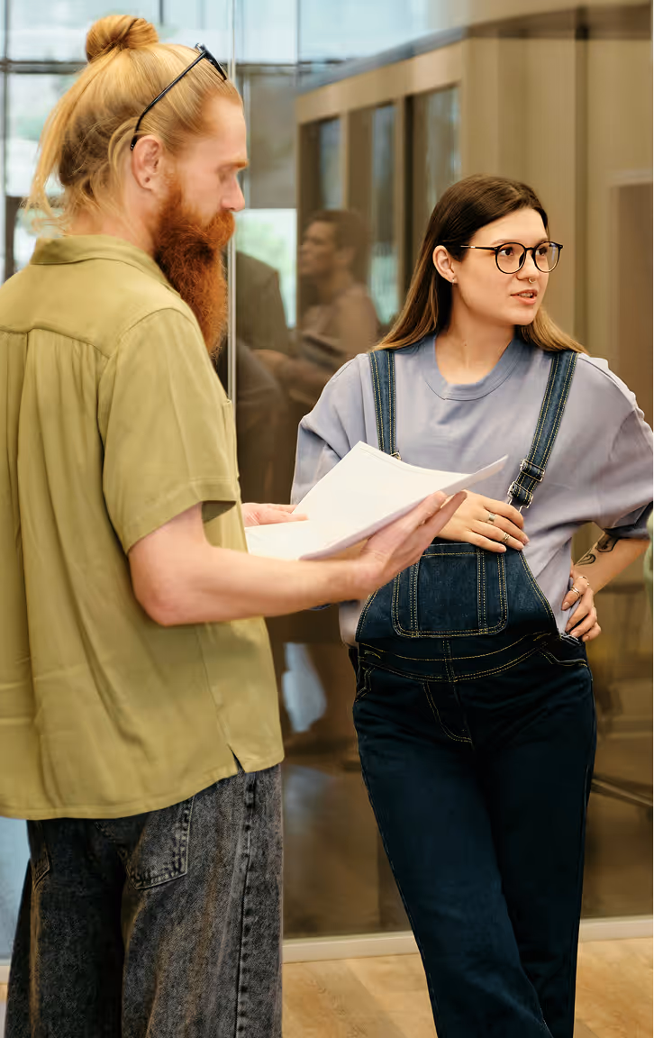 Un homme avec une barbe et un chignon lit des documents pour une femme aux lunettes en salopette.