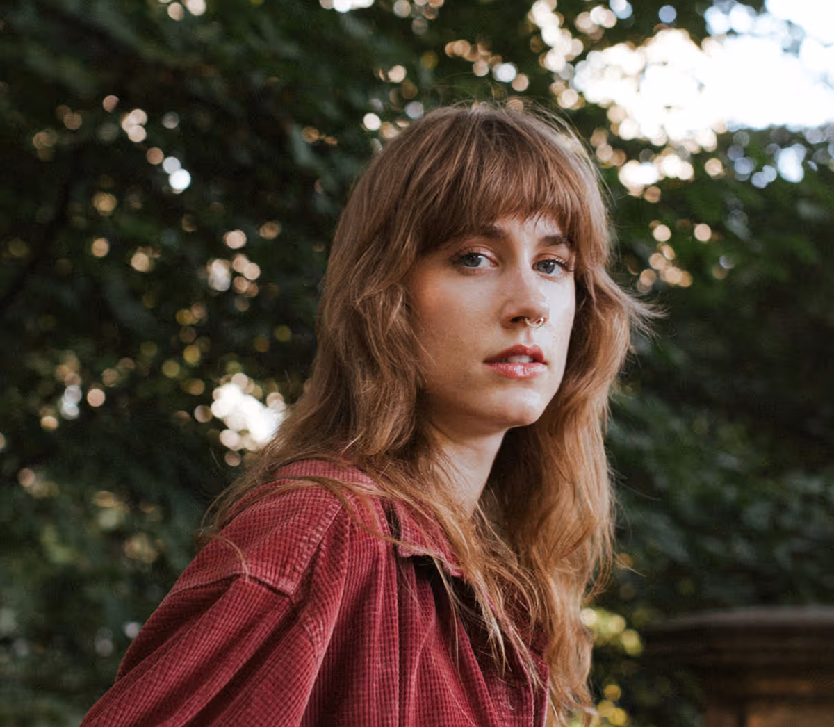Femme aux cheveux longs et roux portant une chemise rouge dans un cadre extérieur naturel.