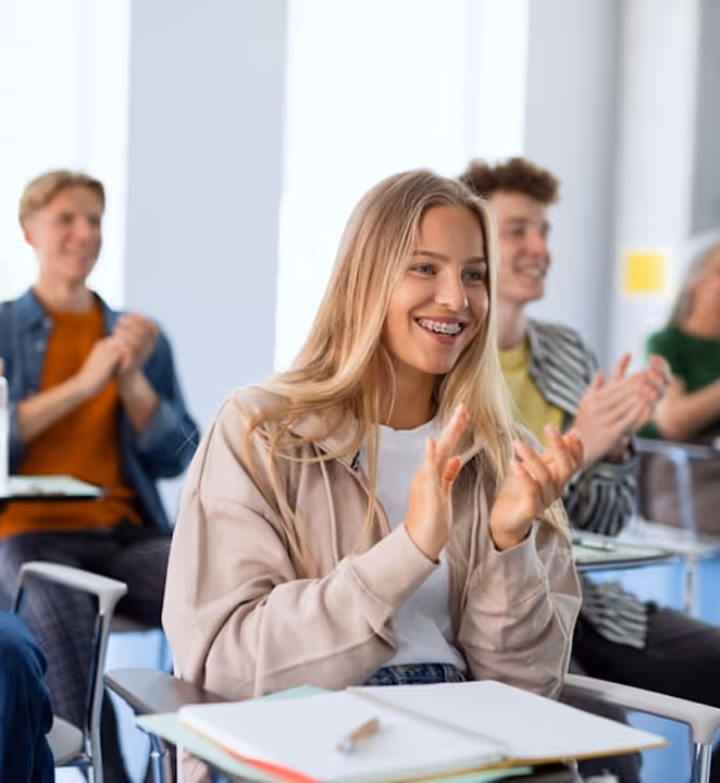 Jeune femme souriante applaudissant en classe avec d'autres étudiants flous en arrière-plan.