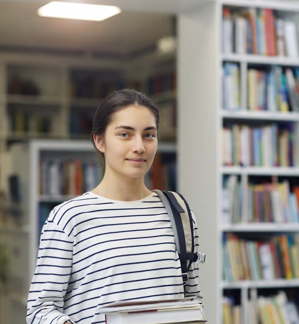 Jeune femme tenant des livres dans une bibliothèque avec un sac à dos à l'épaule.