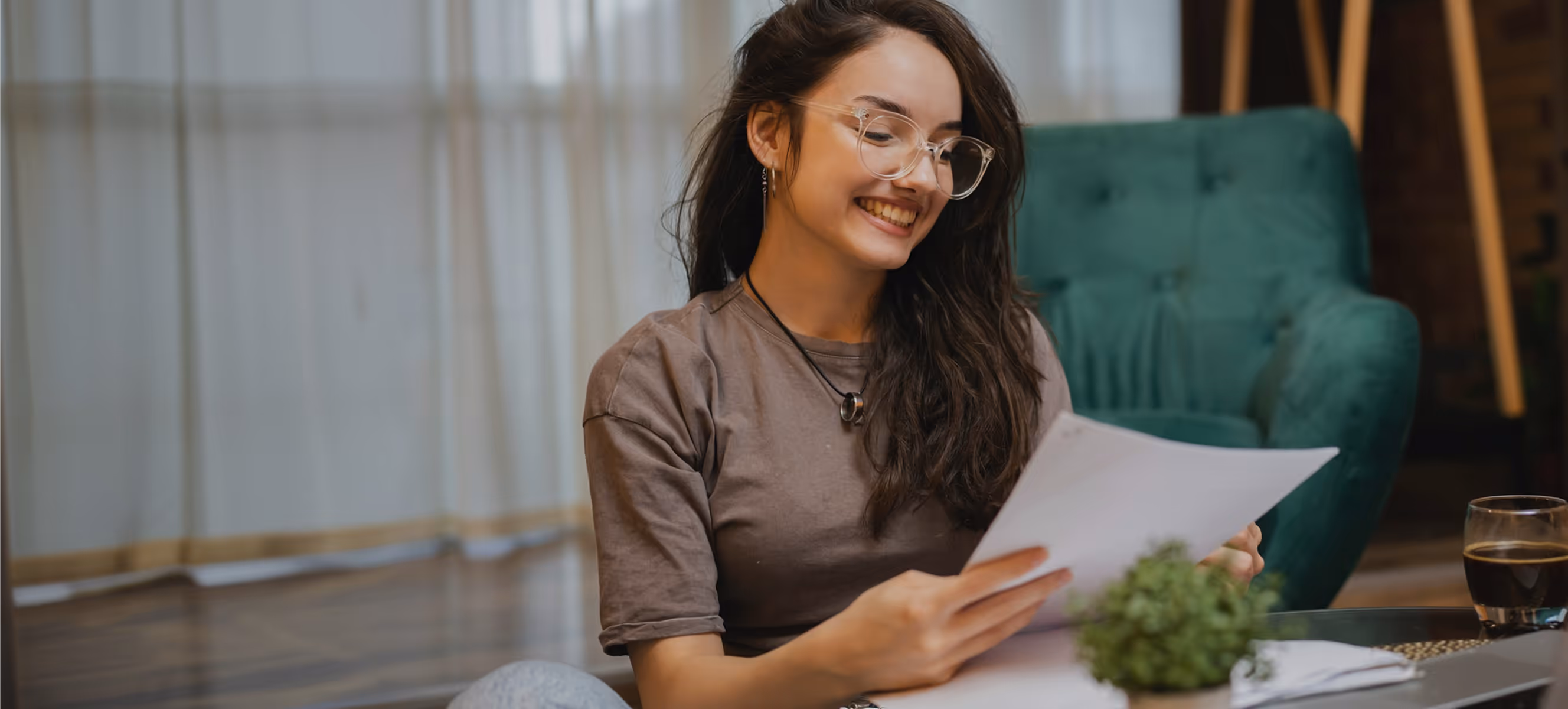 Jeune femme souriante lisant un document assise à la maison, avec une plante et une boisson à côté.