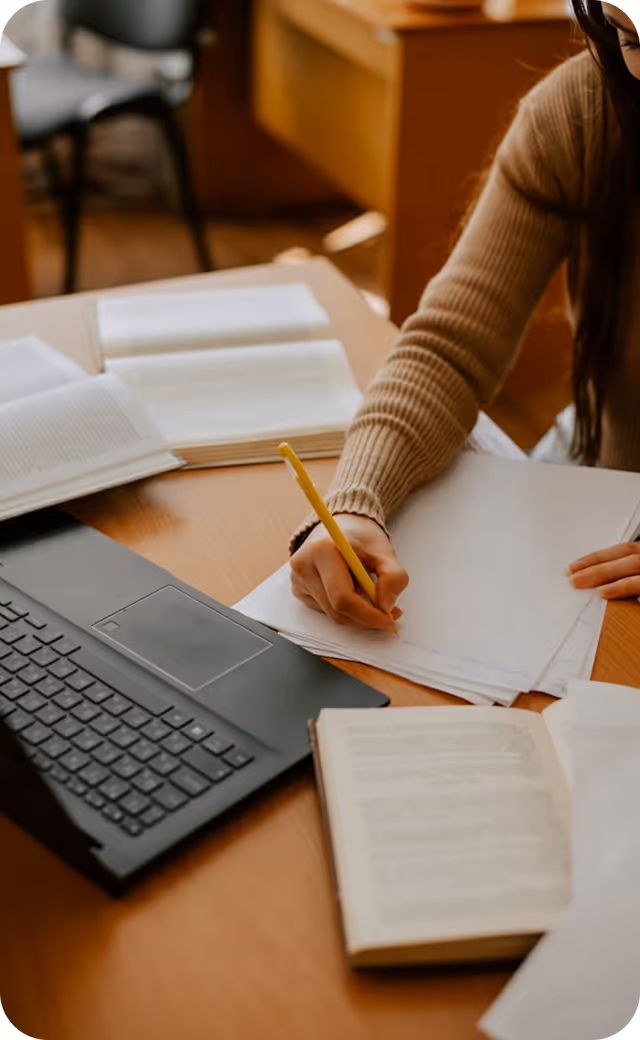 Une personne écrit sur des feuilles blanches avec un crayon près d'un ordinateur portable et des livres ouverts.