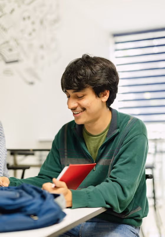 Jeune homme souriant en pull vert tenant un carnet rouge assis à une table d'étude.
