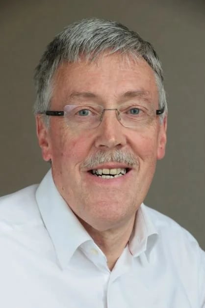Smiling middle-aged man with glasses, gray hair, and a mustache wearing a white shirt against a plain background.