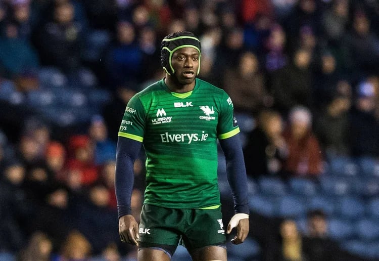 Rugby player in green jersey and headgear standing on field with blurred crowd behind.