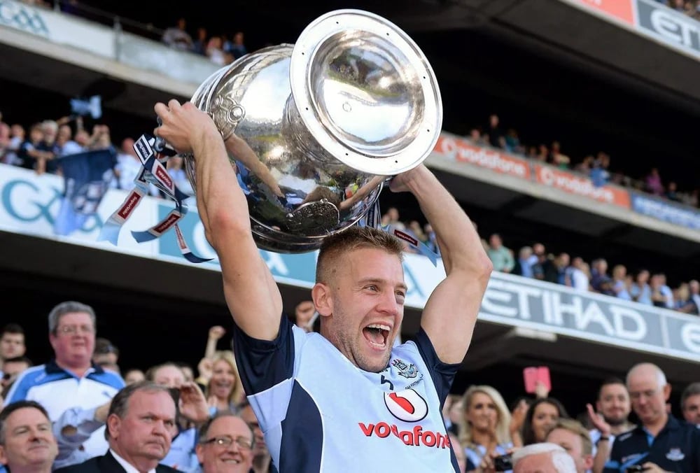 Man in sports jersey lifting a large silver trophy jubilantly in front of a cheering crowd.