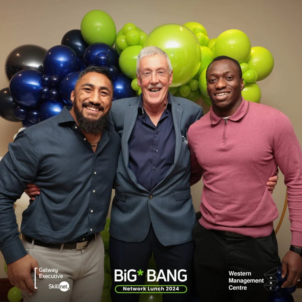 Three men smiling with arms around each other in front of green and blue balloons at network lunch.