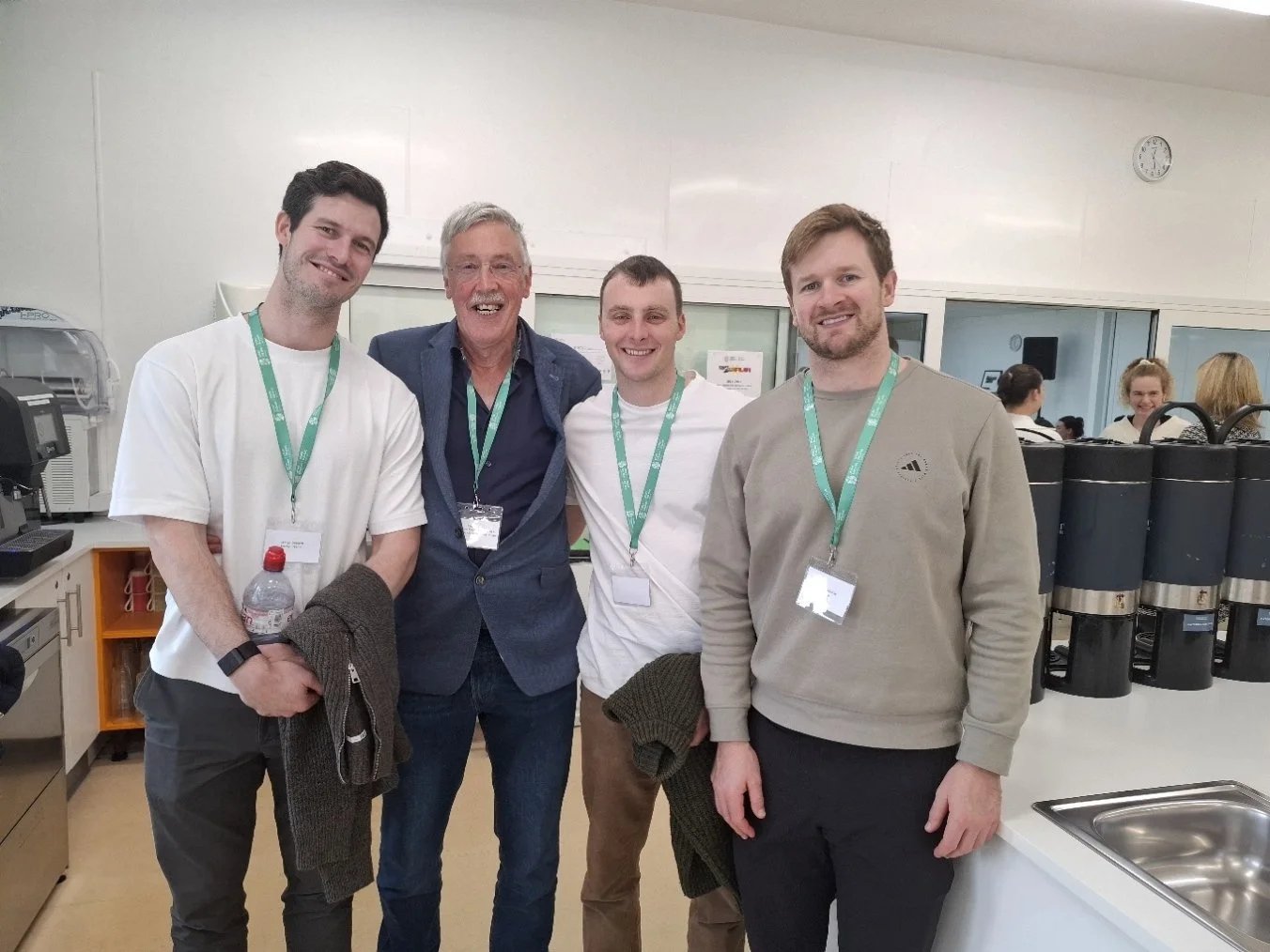 Four men with green lanyards smiling in a kitchen area with coffee urns and a sink.
