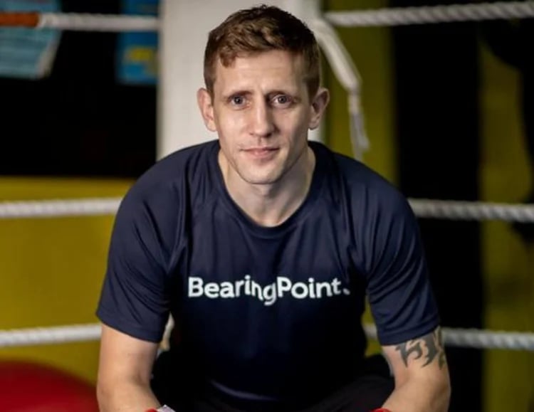 Man wearing a BearingPoint shirt sitting inside a boxing ring with gloves on his hands.