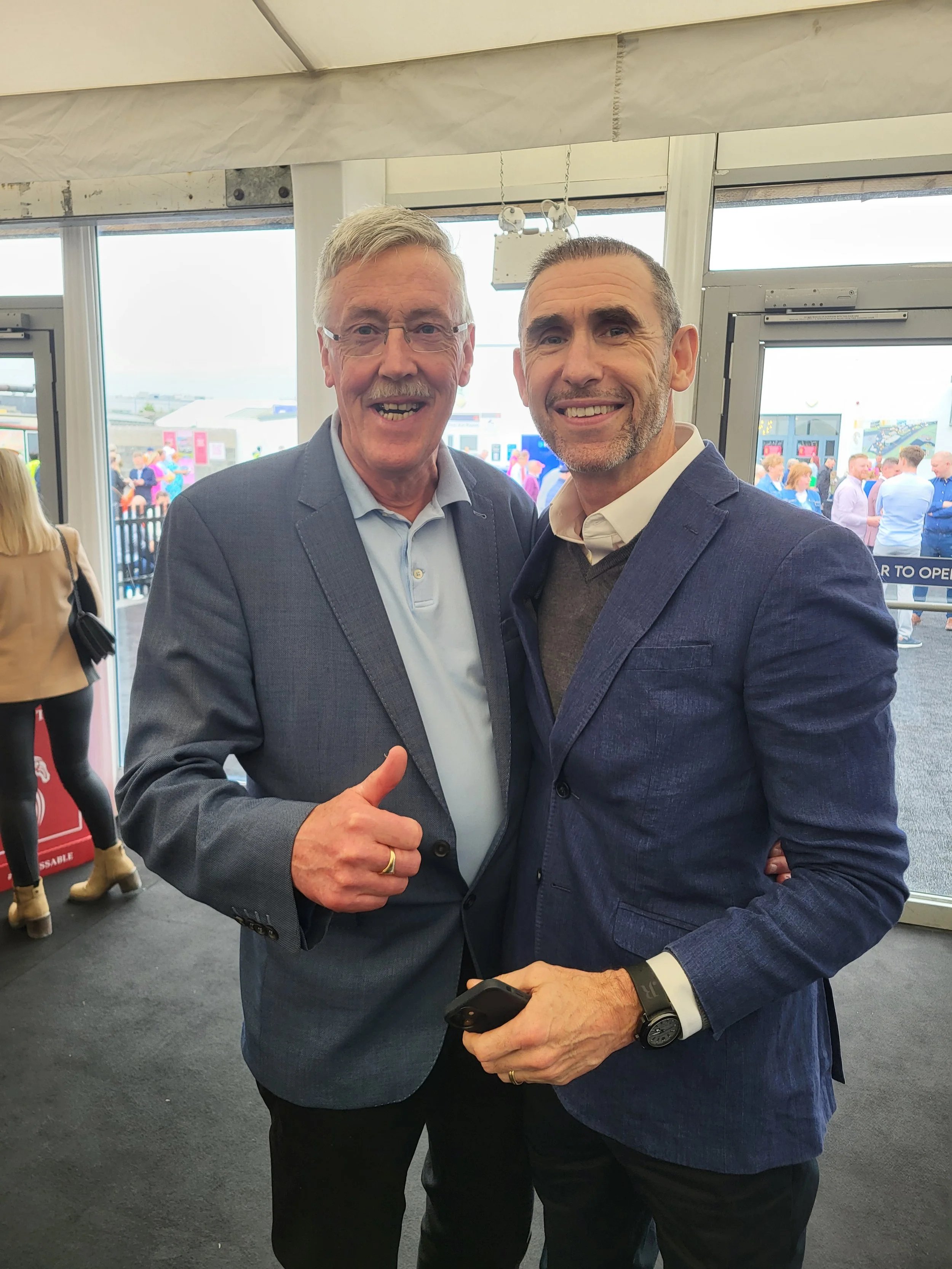 Two men wearing blazers smile at camera; one gives a thumbs-up in a crowded indoor event space.