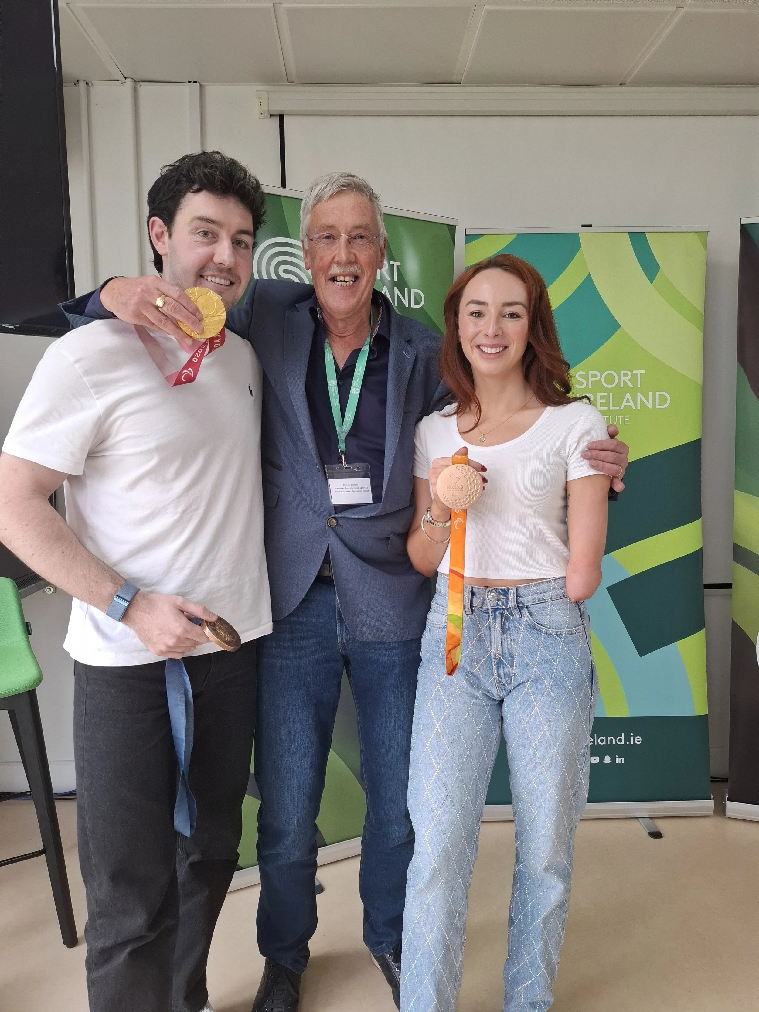 Three people smile at the camera, two holding Olympic medals, standing in front of Sport Ireland banners.