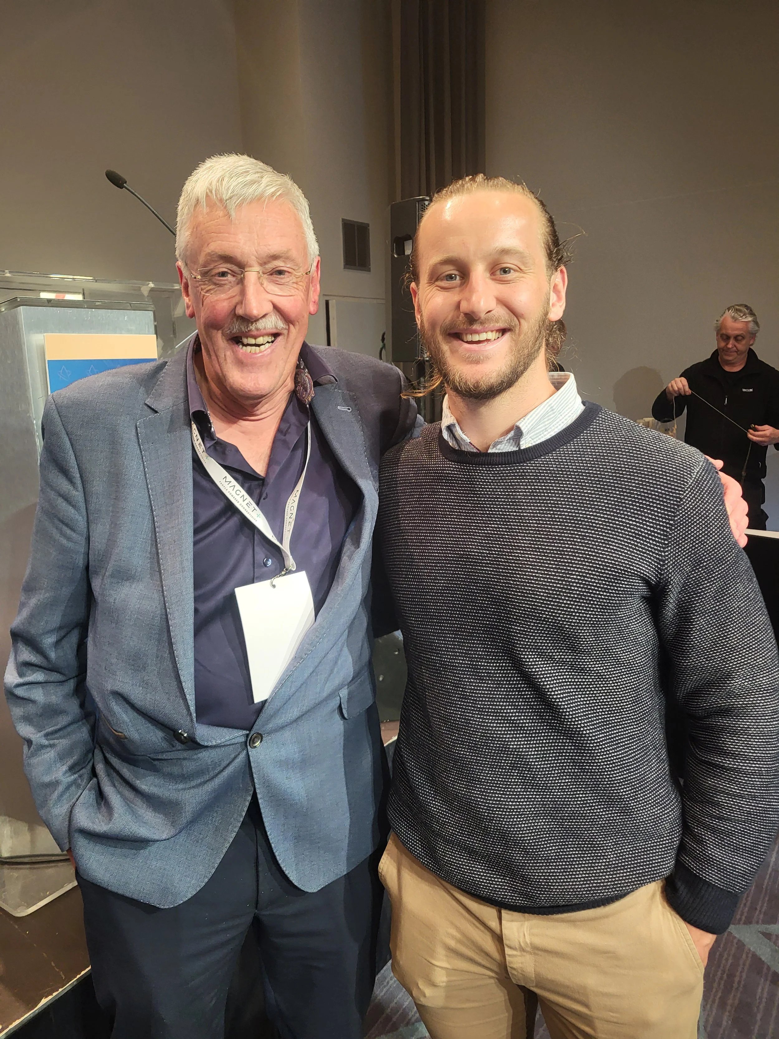 Two men smiling, one older with glasses and blazer, one younger in sweater and khaki pants.