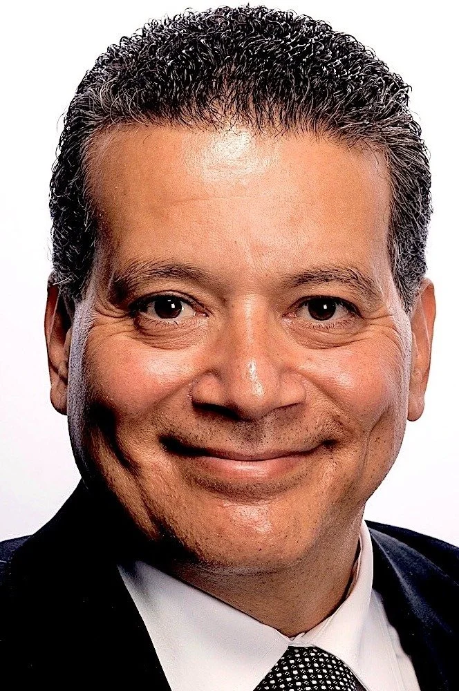Man with short curly hair and a suit smiling at the camera with a white background.