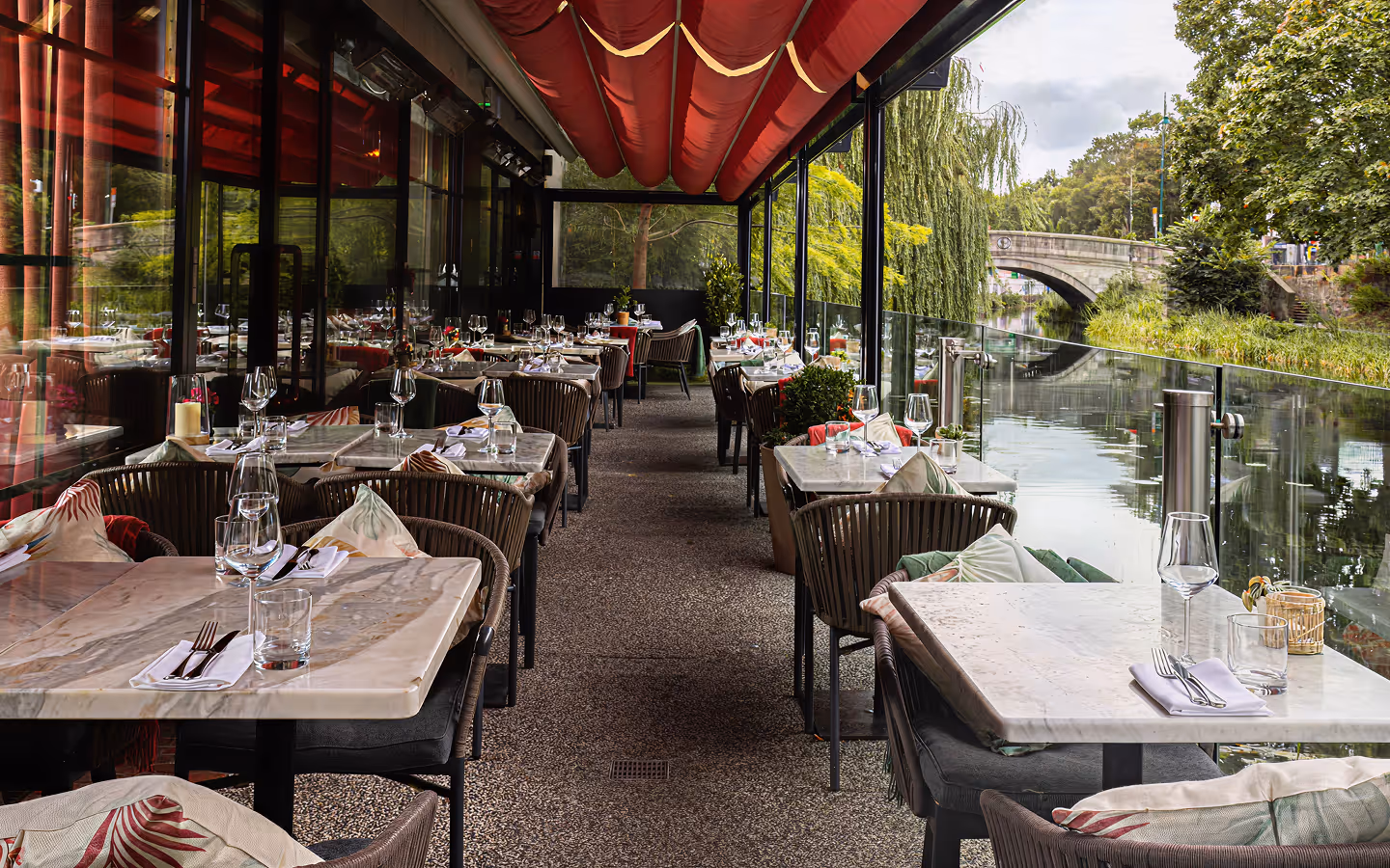 Marble tables under a red awning on the canalside terrace, with a stone bridge and willows beyond, at Angelina's
