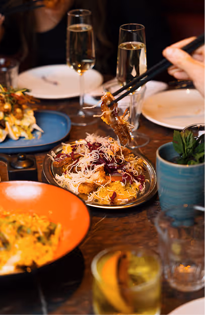 Chopsticks lifting a bite from shared plates alongside cocktails at Mama Yo