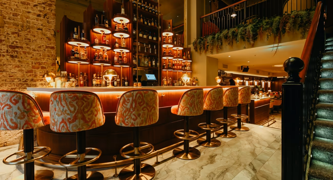 Patterned swivel stools along a curved timber bar with lit spirit shelves and exposed brick