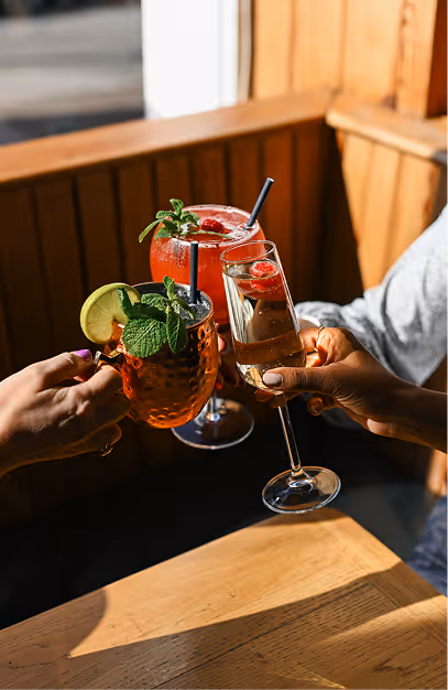 Hands toasting a copper mule mug, a berry cocktail and a sparkling wine flute in a timber booth at Elephant & Castle