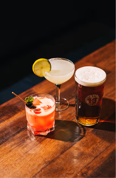 Two cocktails and a pint of red ale on a sunlit wooden table at Captain Americas