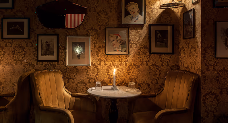 Velvet armchairs and a candlelit marble table amid framed prints at Vintage Cocktail Club