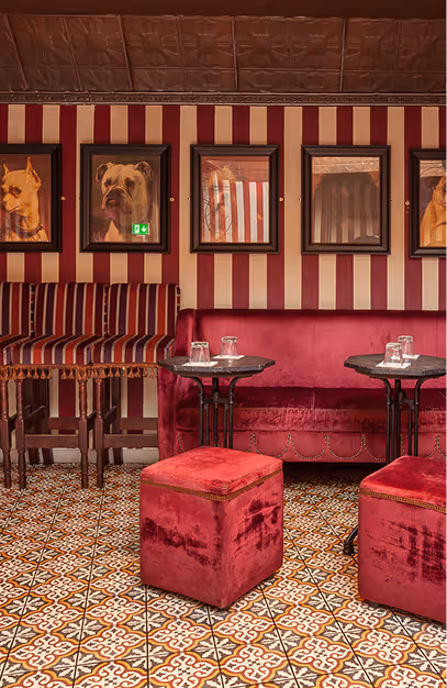 Framed dog portraits above a pink velvet banquette and red ottomans at Vintage Cocktail Club