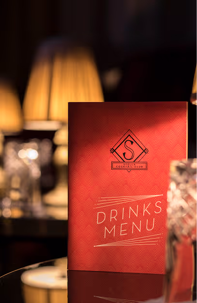 Art deco drinks menu on a lamp-lit table at Stella Cocktail Club