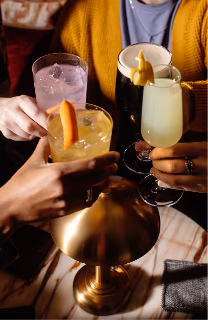 Four hands raising cocktails over a marble table beside a brass lamp at The Stapleton