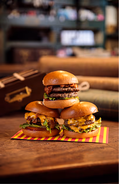 Three stacked cheeseburgers on striped paper on a wooden table at John's Bar & Haberdashery