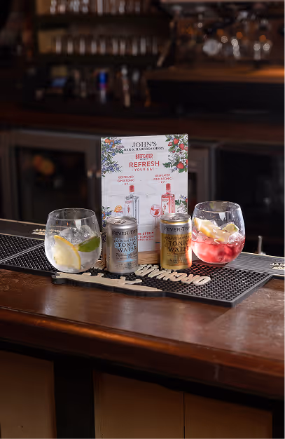 Two gin and tonics with tonic cans and a promo card on the bar at John's Bar & Haberdashery