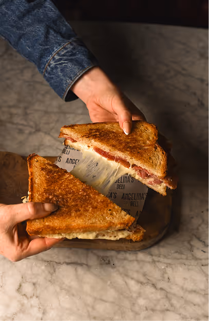Hands lifting a halved cheese toastie off branded paper on a wooden board at Angelina's Deli