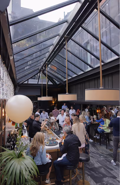 Busy glass-roofed atrium bar with guests at the counter under drum pendants at The Grayson