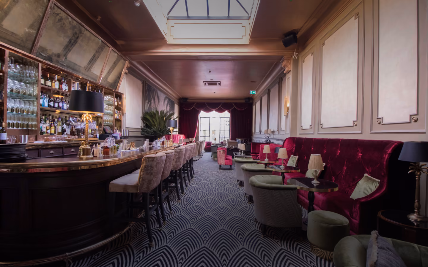 Curved brass bar and stools facing a raspberry velvet banquette on Art Deco carpet at Stella Cocktail Club