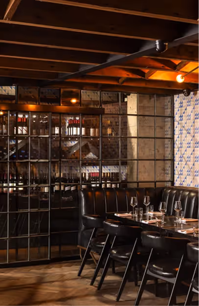 Glass-fronted wine wall beside a black leather banquette and set tables under timber beams at Angelina's