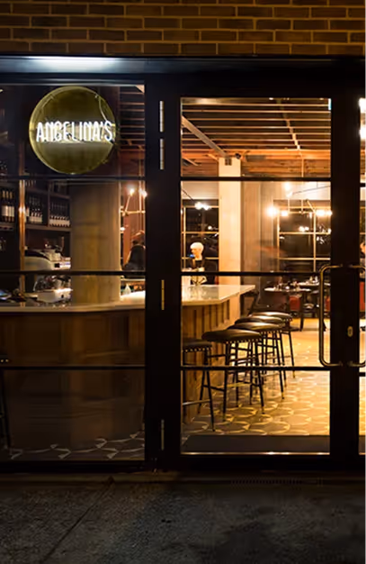 Lit green Angelina's sign above black-framed glass doors, with the bar and stools visible inside at night