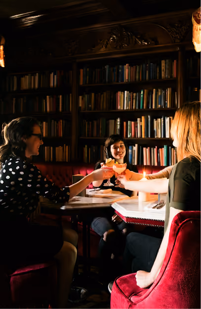 Three colleagues toasting cocktails in a wood-panelled bar lined with bookshelves
