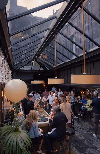 Guests at a busy central bar under a glass atrium roof during a private party