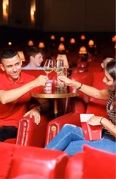 Couple toasting white wine over popcorn in red leather recliner seats at Stella Cinema