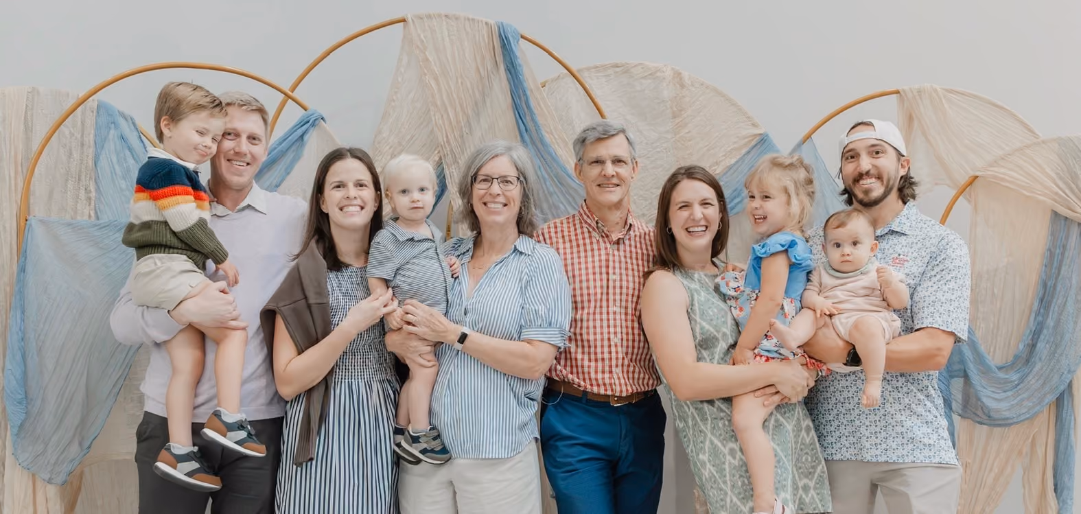 A smiling multi-generation family posing indoors with young children and decorative fabric in the background.