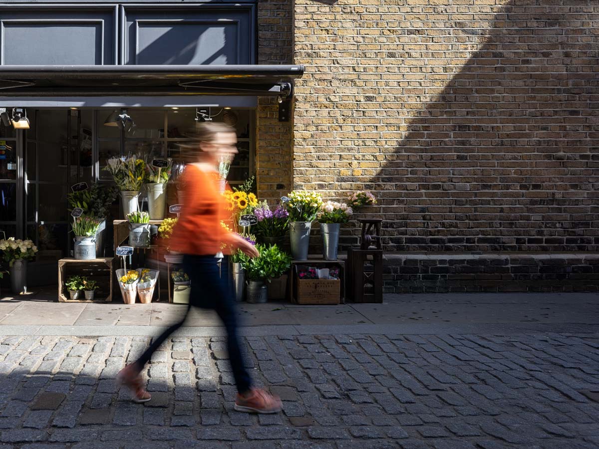 Blurred person walking past a flower shop with bouquets displayed outside on a sunny cobblestone street.