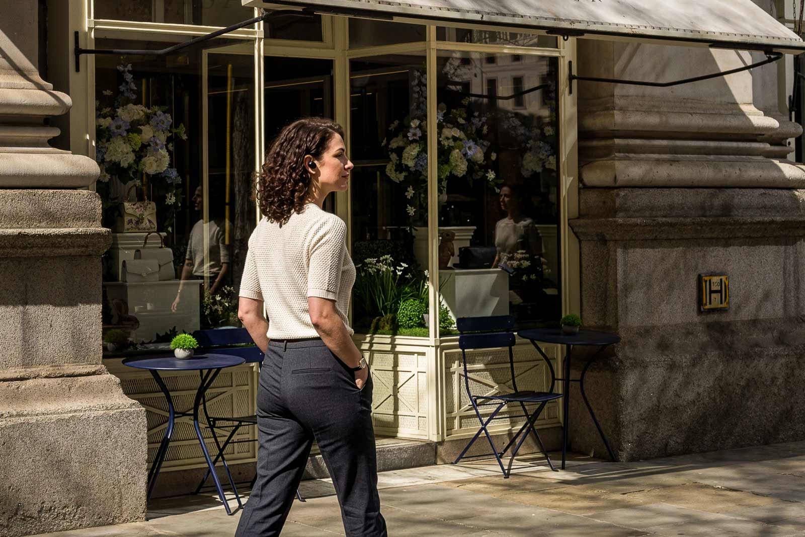 Woman with curly hair wearing earbuds and light sweater walking past a café with outdoor tables.