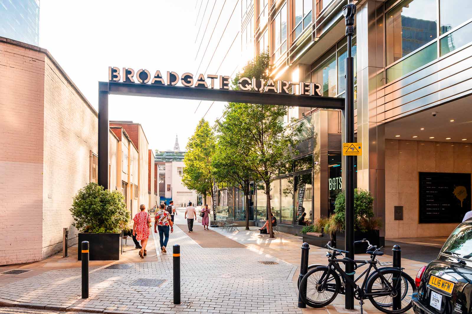 Street entrance to Broadgate Quarter with people walking, trees, a bike, and parked car.