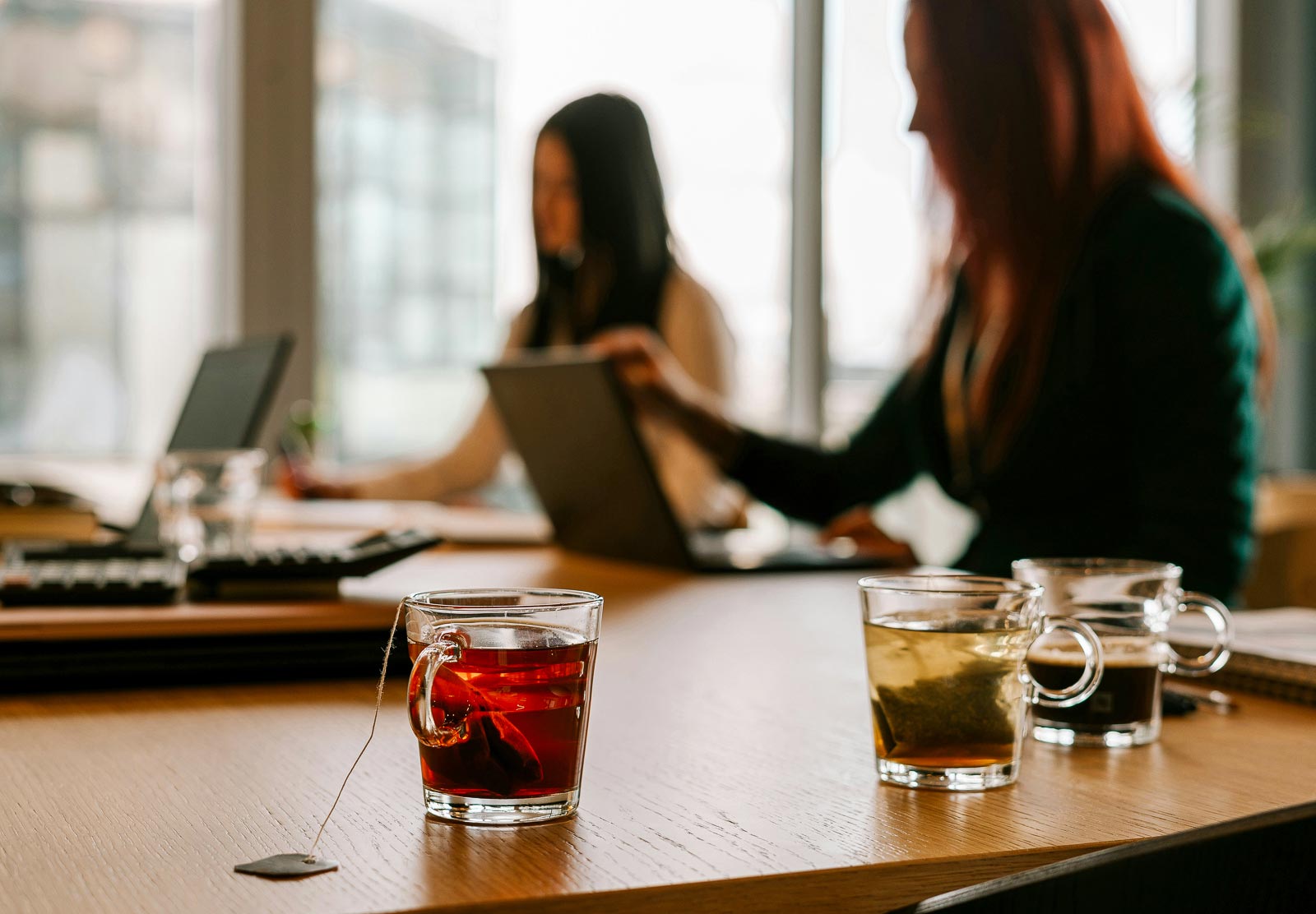 Three glass cups with different teas on a table with blurred people working on laptops in the background.