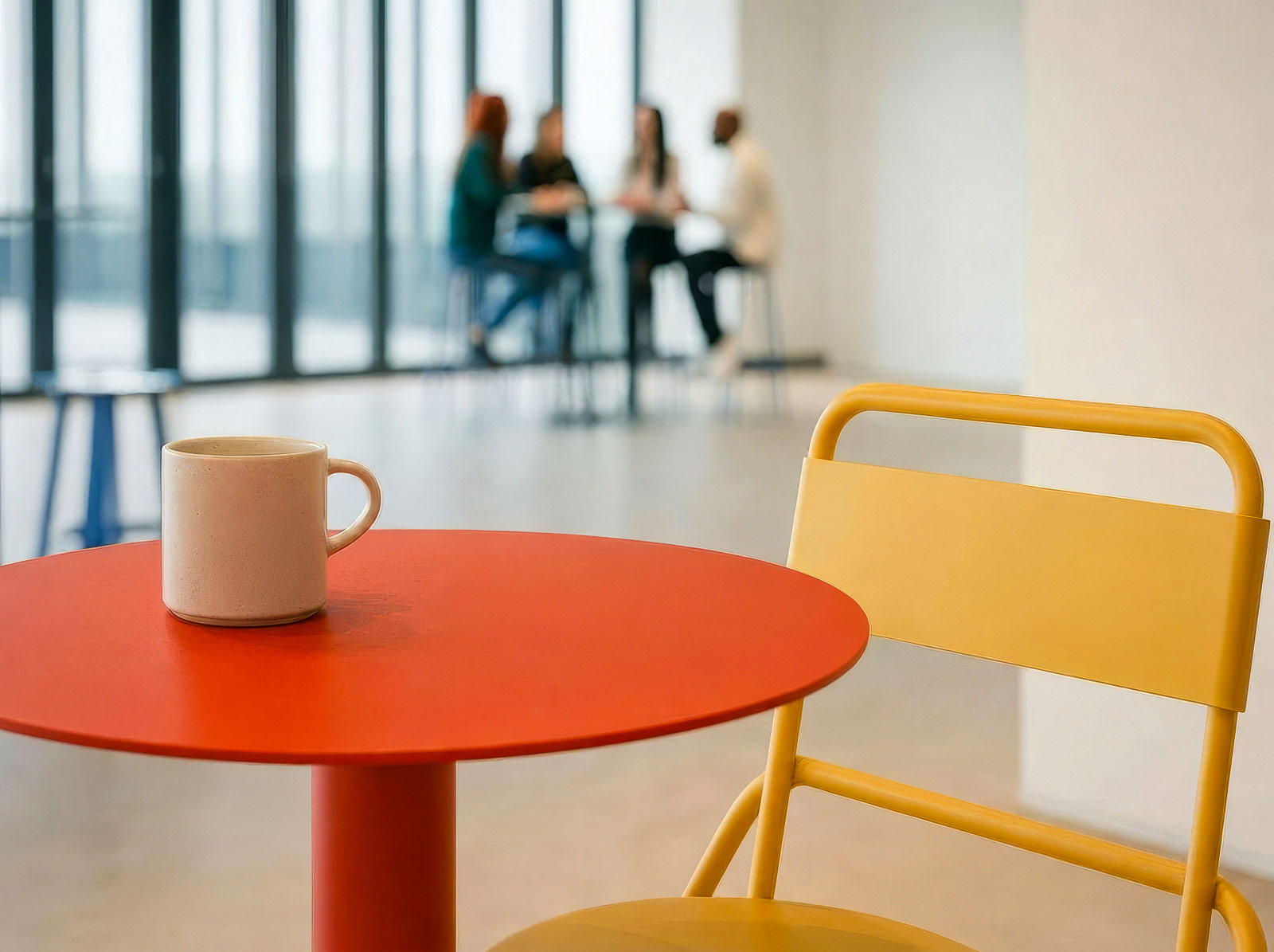 A ceramic mug on an orange round table with a yellow chair in a bright office with people in the background.
