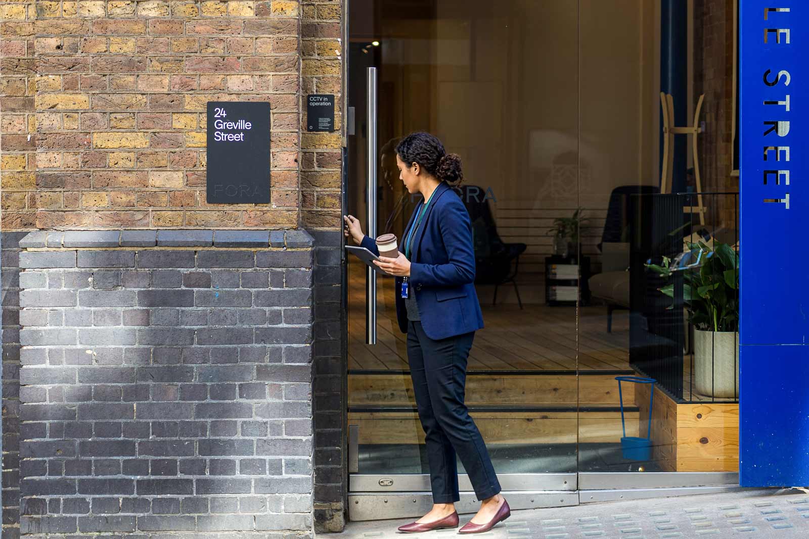 Woman holding coffee and tablet unlocking a glass door at 24 Greville Street entrance, London.