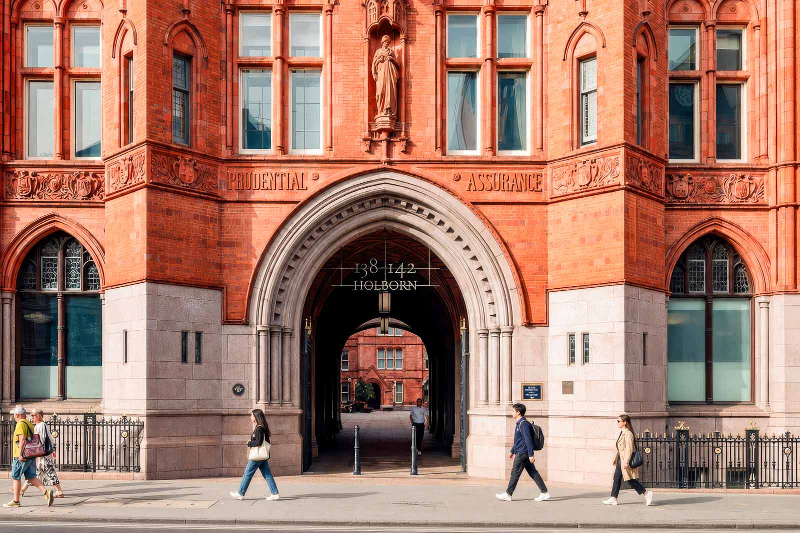 People walking past the detailed red brick archway entrance of the Prudential Assurance building on Holborn.