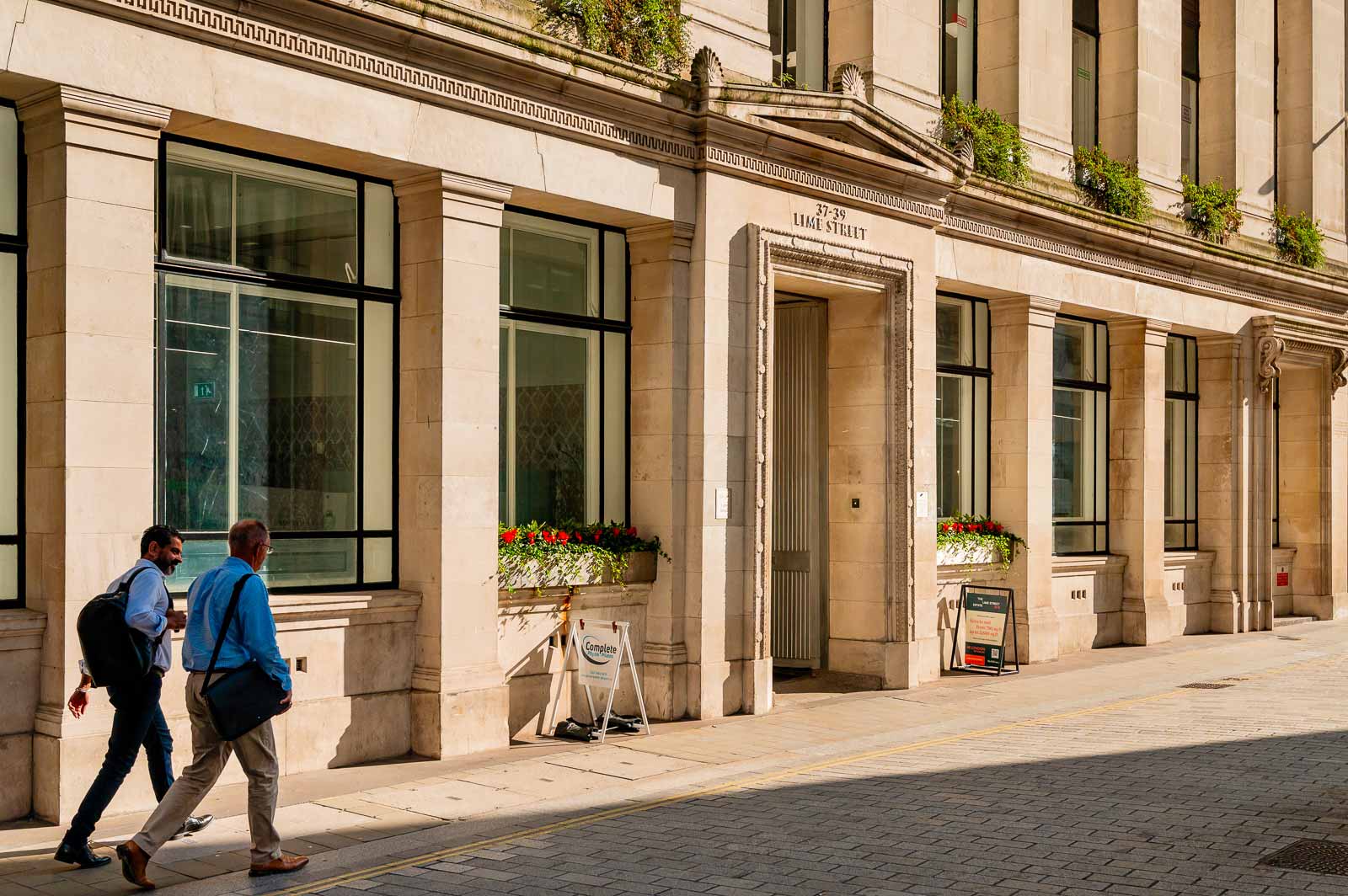 Two men walking past a beige stone building with large windows and flower boxes on a sunny London street.
