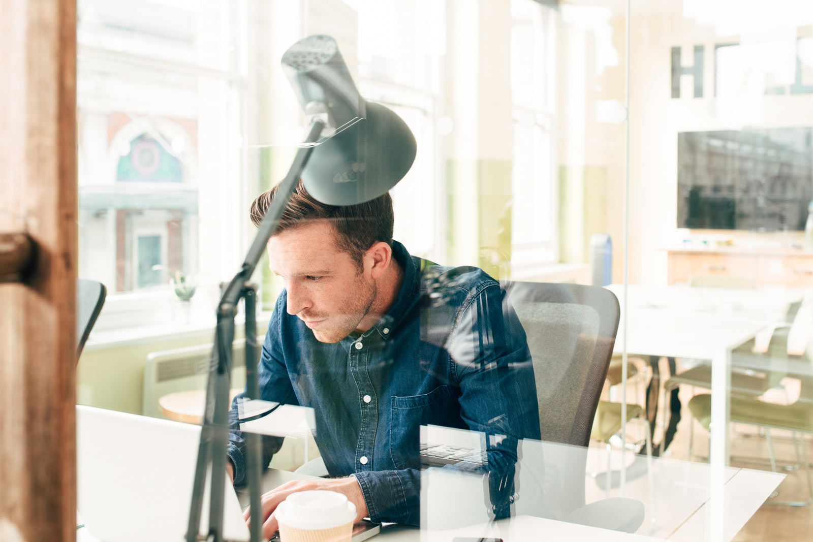 Man in denim shirt working on laptop at a desk in a bright office with glass walls.