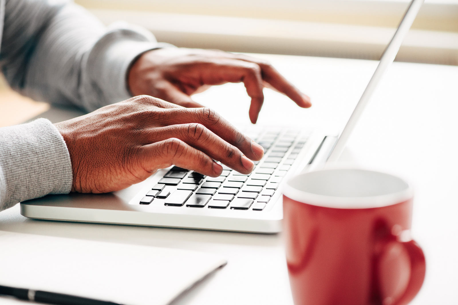Somebody working, a close-up of hands typing on a laptop keyboard with a red coffee mug and pen on a white desk.